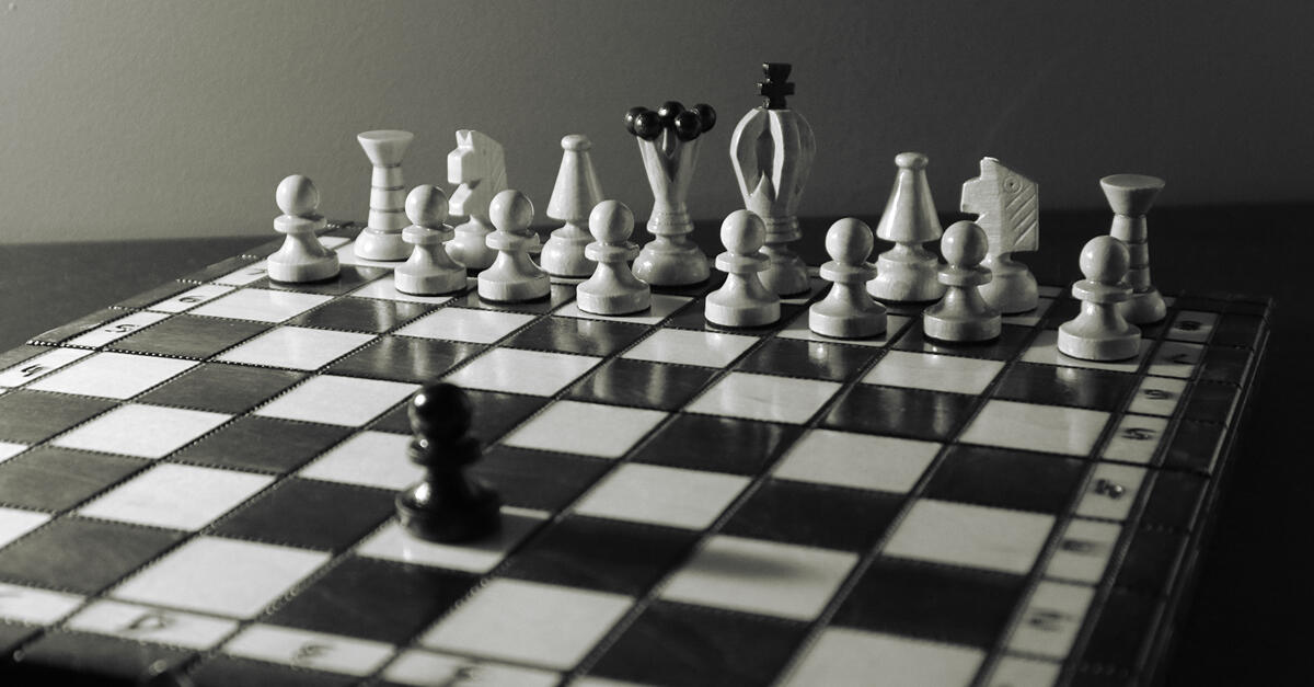 A black pawn stands alone at the center of a chessboard, facing a full line of white pieces arranged in perfect formation. The image is in grayscale, emphasizing the stark contrast and tension—symbolizing isolation, imbalance, and the quiet weight of navig