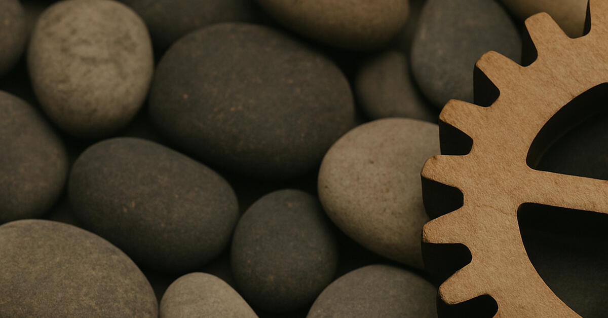 Close-up image of smooth, rounded river stones in varying shades of gray and brown. A single wooden gear appears partially in the bottom right corner, contrasting the organic texture of the stones with the structured shape of the gear—symbolizing the tensi