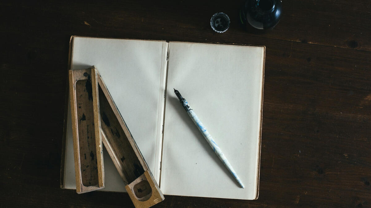 Every blank page begins with intention, not certainty. An open blank notebook lies on a wooden desk beside a marble-handled dip pen, an ink bottle, and a wooden pen case, evoking the start of a creative or reflective process.