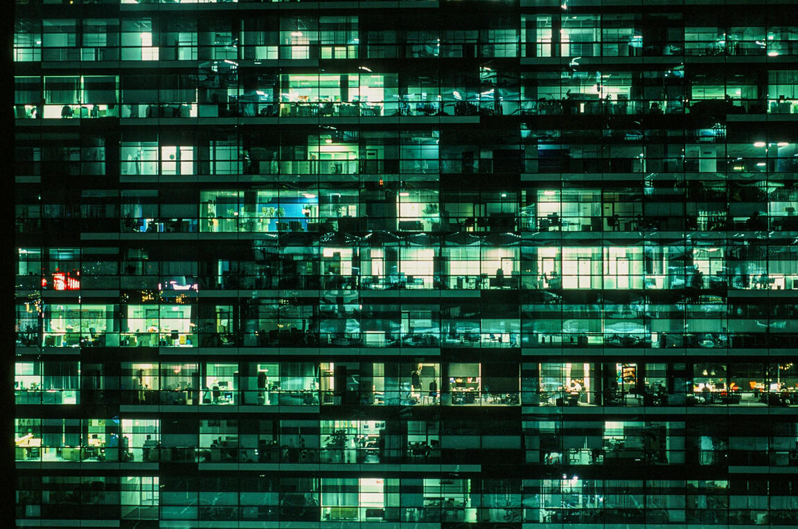 Every window a workflow. Every light a node in a living network. A nighttime view of a glass office building with dozens of lit windows revealing people working inside, symbolizing systems and human structure.