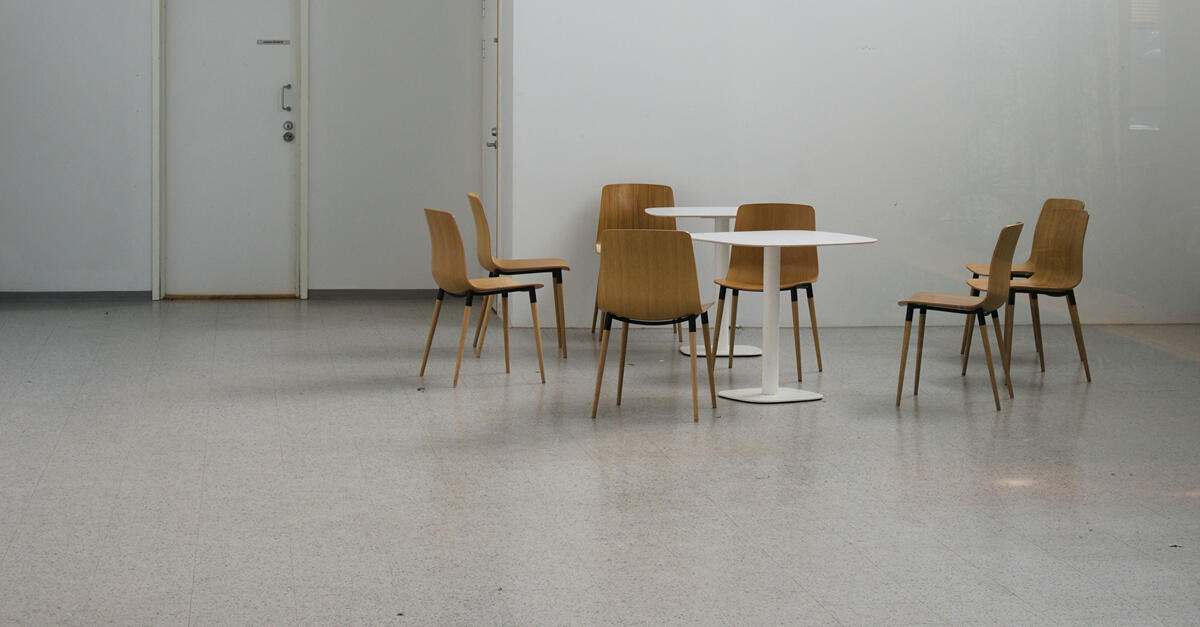A minimalist scene of two small round tables surrounded by several wooden chairs in an empty room with a plain white wall. One chair is slightly apart from the group, creating a quiet, contemplative sense of absence.