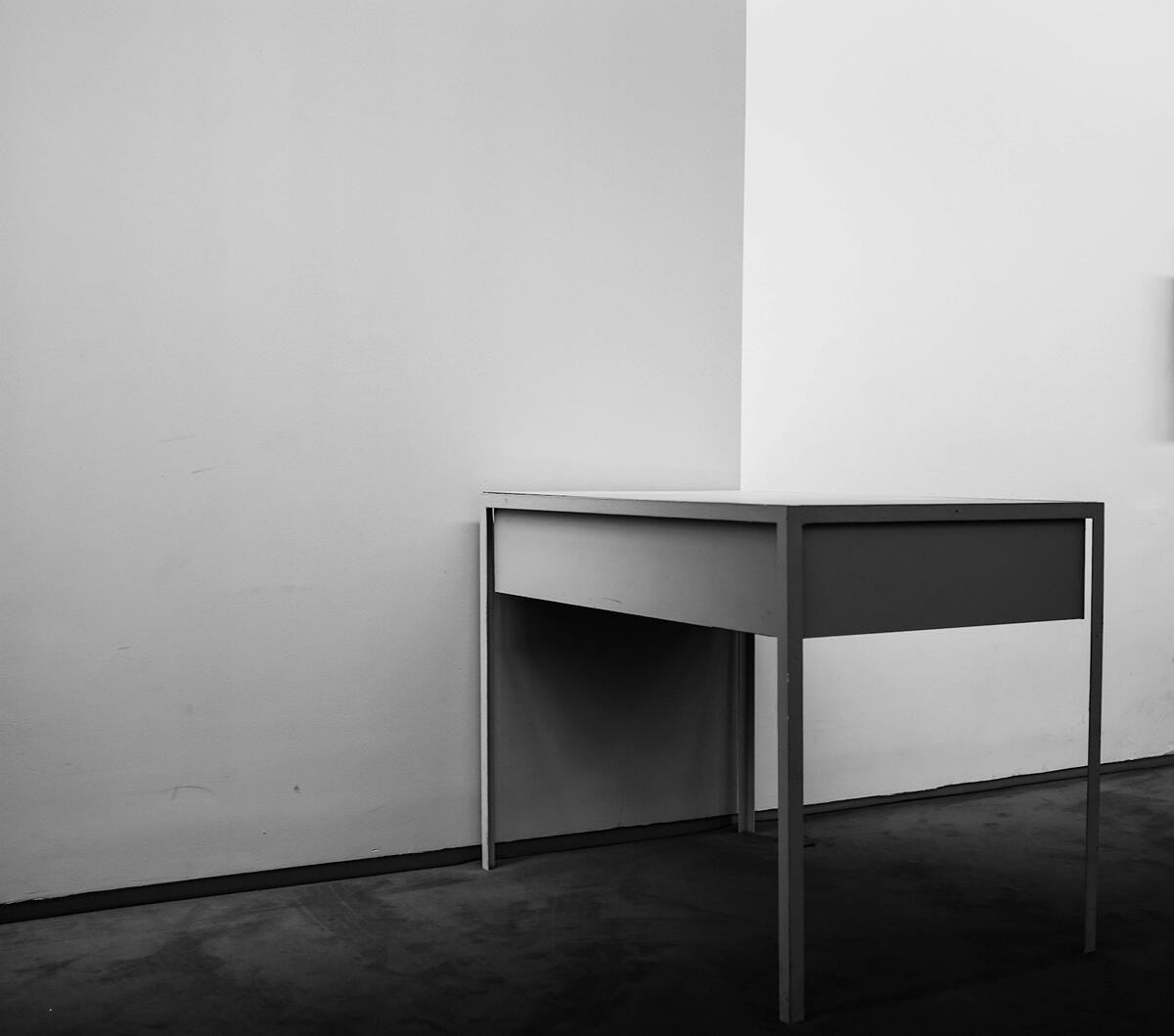 A minimalist scene of two small round tables surrounded by several wooden chairs in an empty room with a plain white wall. One chair is slightly apart from the group, creating a quiet, contemplative sense of absence.