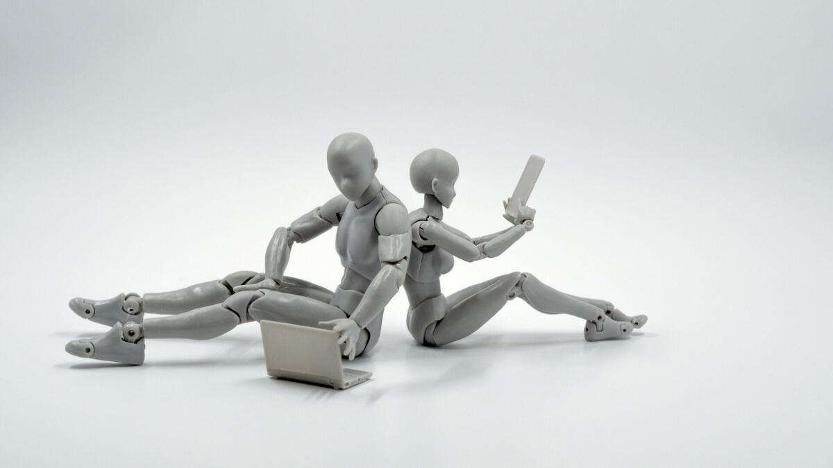 Two gray humanoid figures sit back-to-back against a white background, one using a laptop and the other holding a tablet, symbolizing human-machine collaboration and digital interdependence.