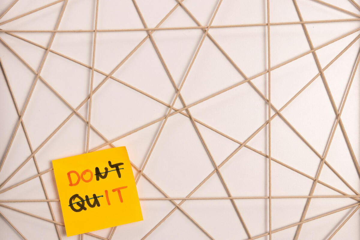 Do it. Don’t quit. A yellow sticky note with the words “DON’T QUIT,” where “DON’T” is crossed out to reveal “DO IT,” set against a geometric web of tan string lines — symbolizing persistence and the interconnected rhythm of effort.