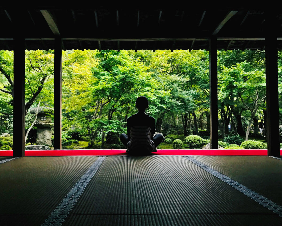 Presence is progress. Person sitting cross-legged at the edge of a temple, gazing out toward a lush green garden — a quiet moment symbolizing patience, presence, and the rhythm of discipline.