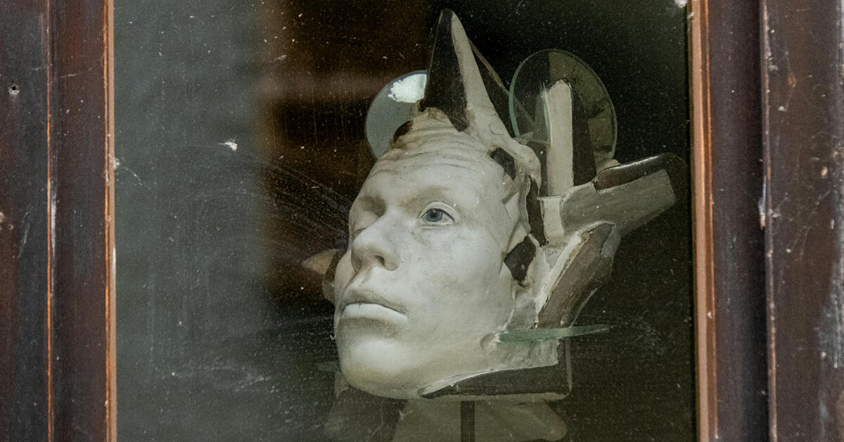 A fragmented ceramic sculpture of a human head displayed behind glass, with visible cracks and protruding pieces suggesting reconstruction or transformation. The expression is calm and introspective, symbolizing the process of self-examination and rebuildi