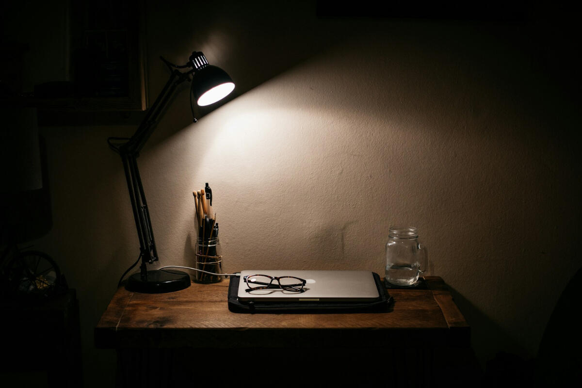 Desk with lamp A dimly lit wooden desk with a black lamp shining on a closed laptop, eyeglasses, and a jar of water — evoking a sense of quiet focus and solitude.